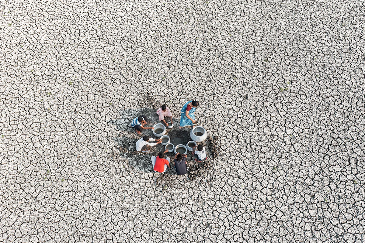 Aerial shot of a group of people with buckets are collecting water from a small source, surrounded by grey cracked earth.
